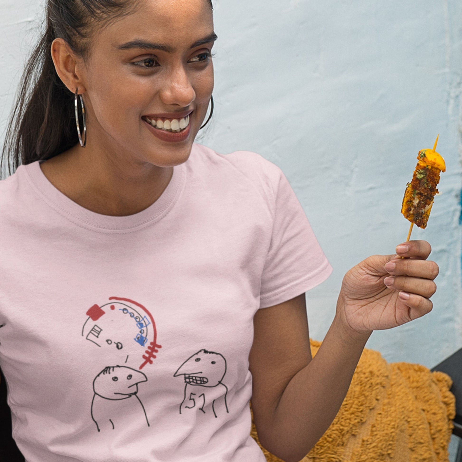 Woman eating some food while wearing a pink t-shirt with a colorful abstract drawing indoors.