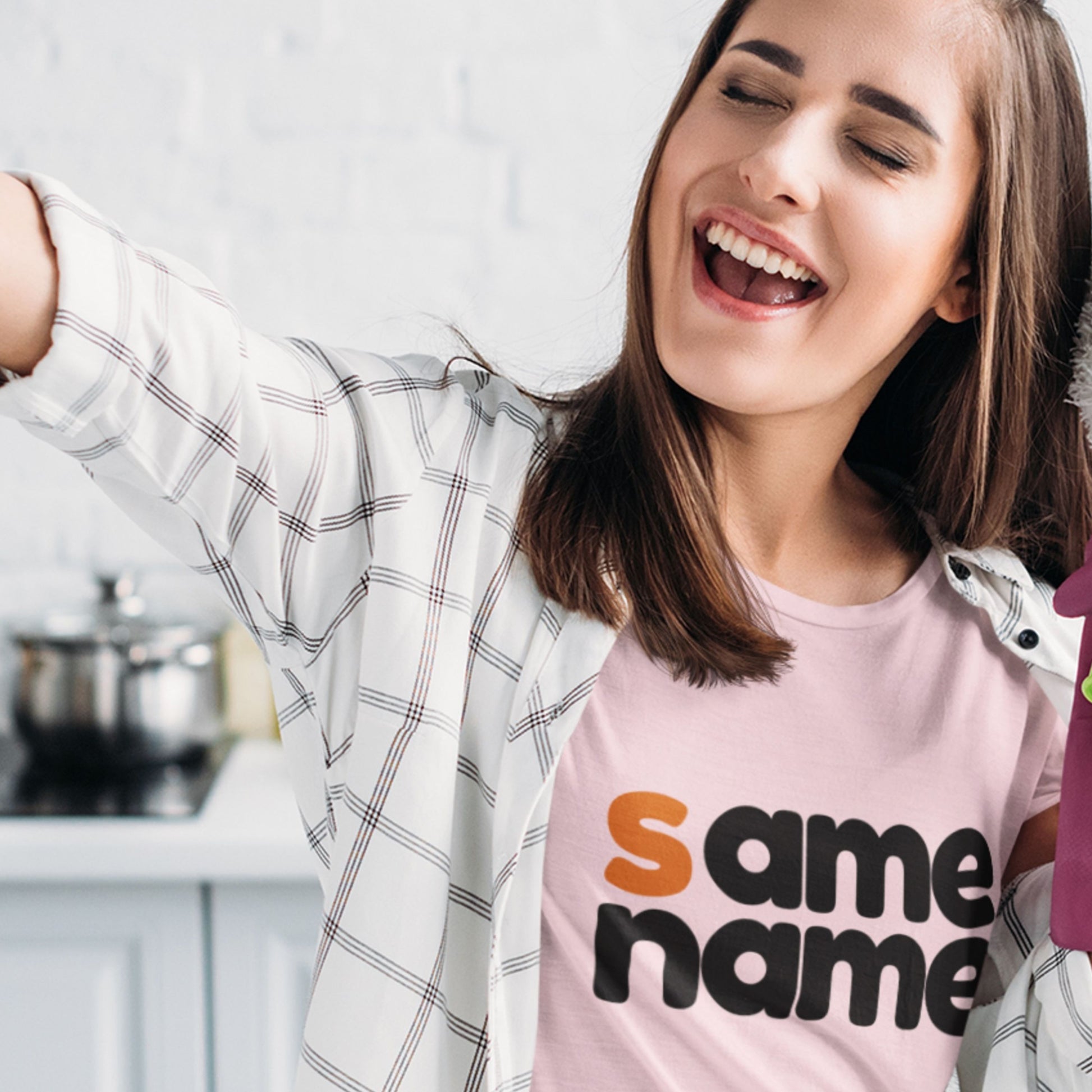 Woman wearing a pink t-shirt with 'same name' text in a kitchen setting.