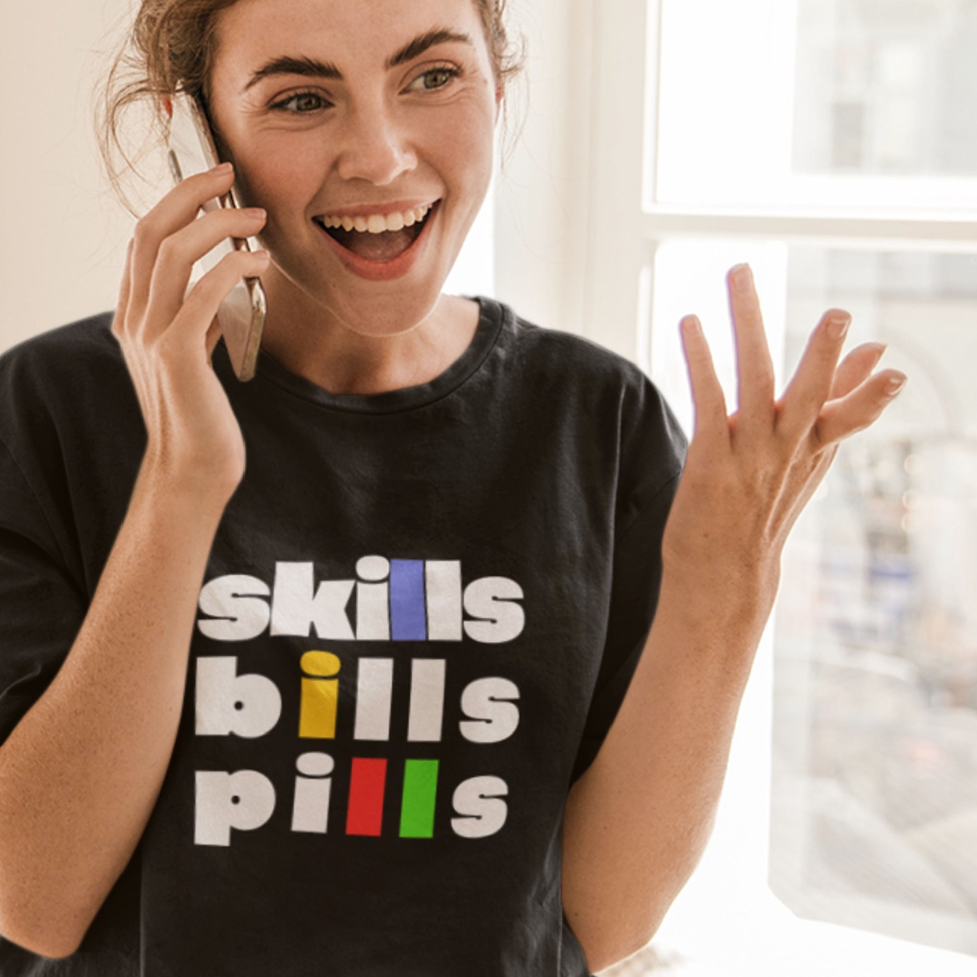 Person wearing a black t-shirt with 'skills bills pills' text, smiling and talking on a phone.