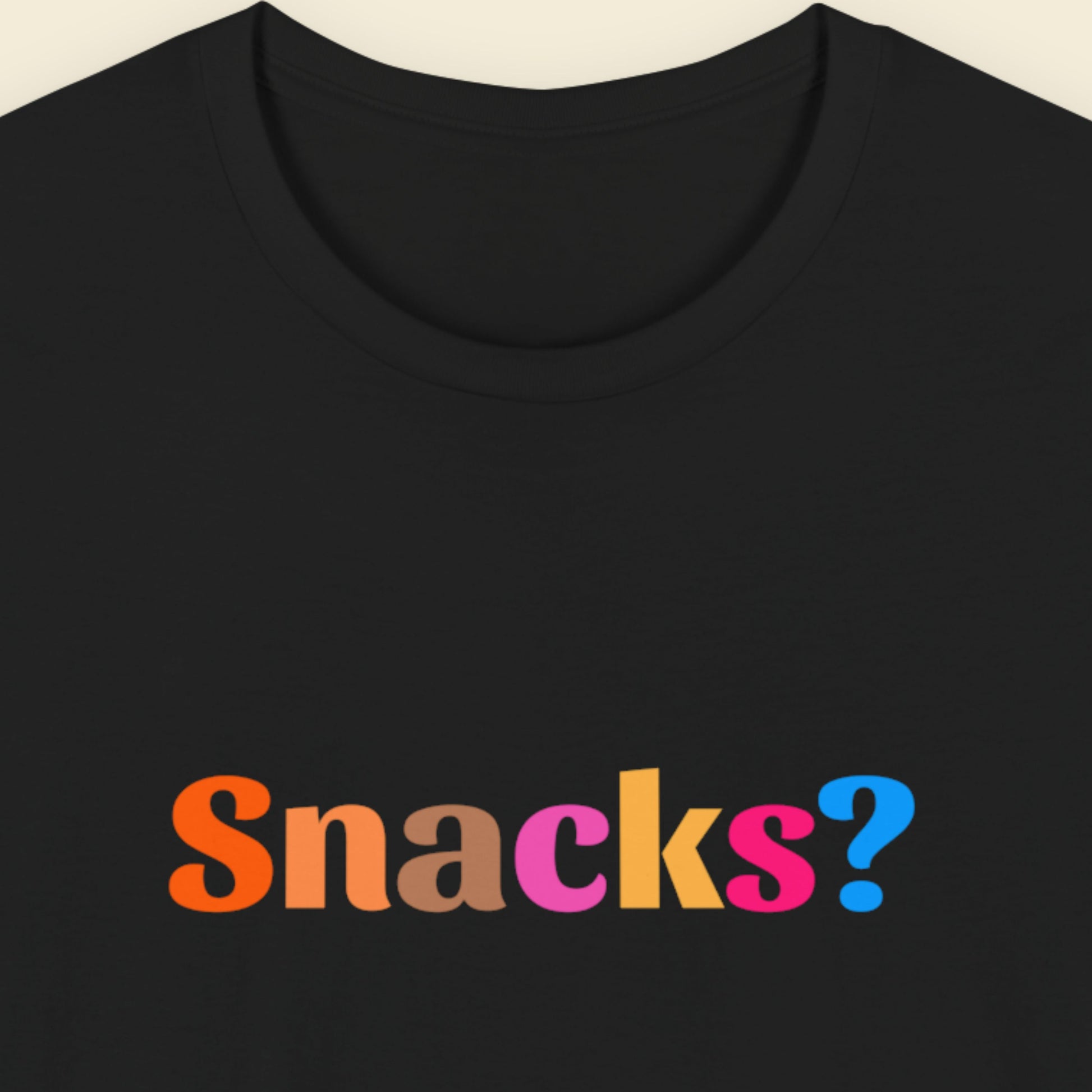 Black t-shirt with 'Snacks?' text in colorful letters on a beige background.