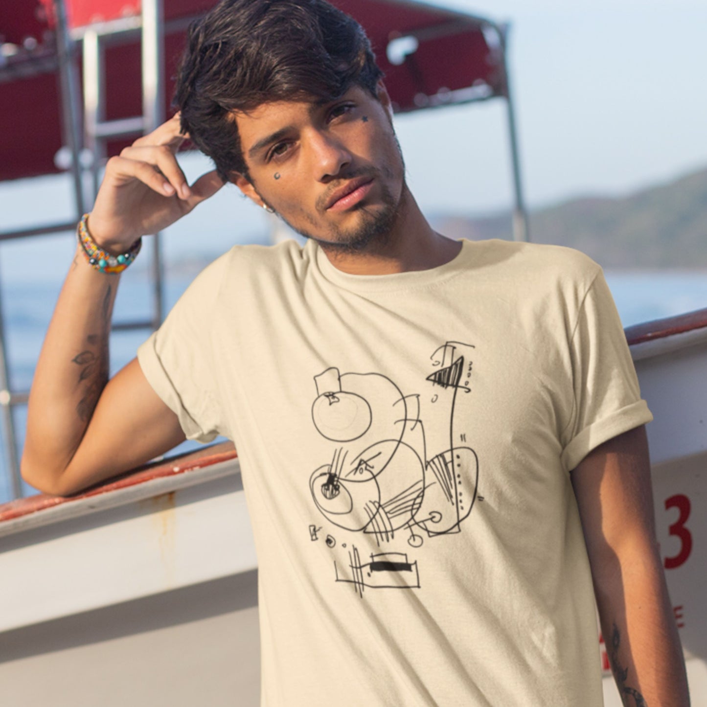 Man wearing a cream colored t-shirt with an abstract line drawing leans on a boat.