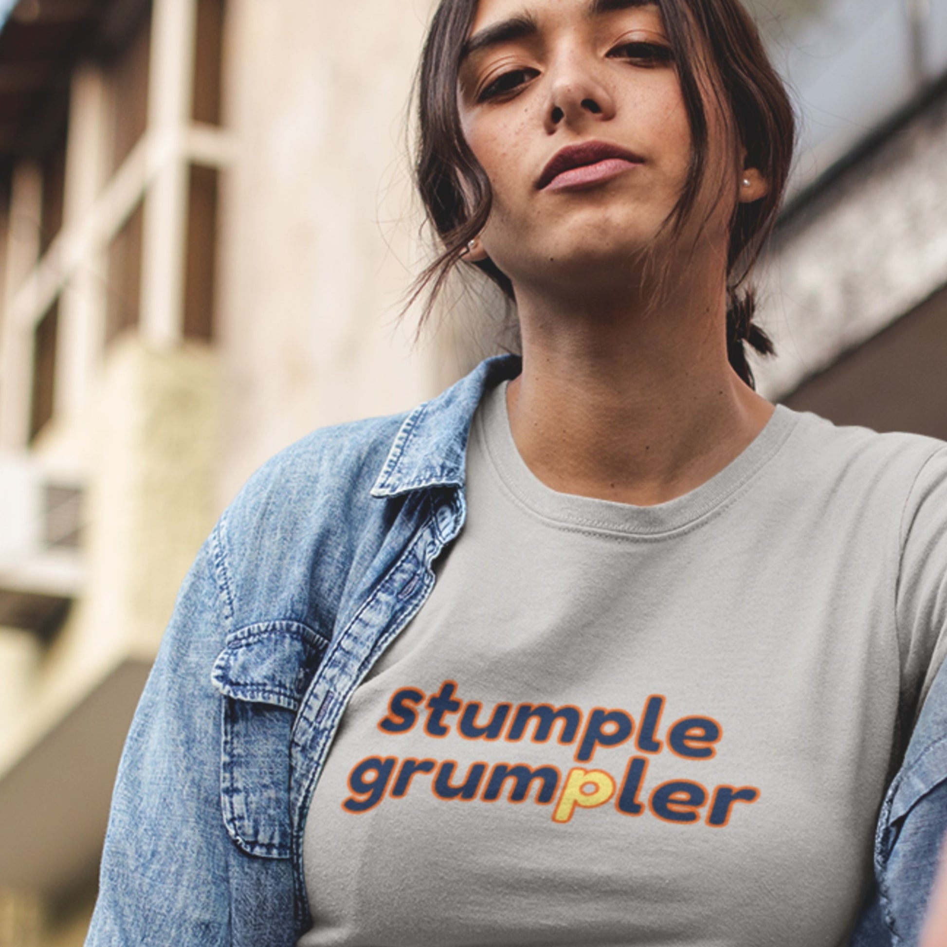 Attitude woman wearing a light grey t-shirt with multicolored 'stumple grumpler' text in front of a building.