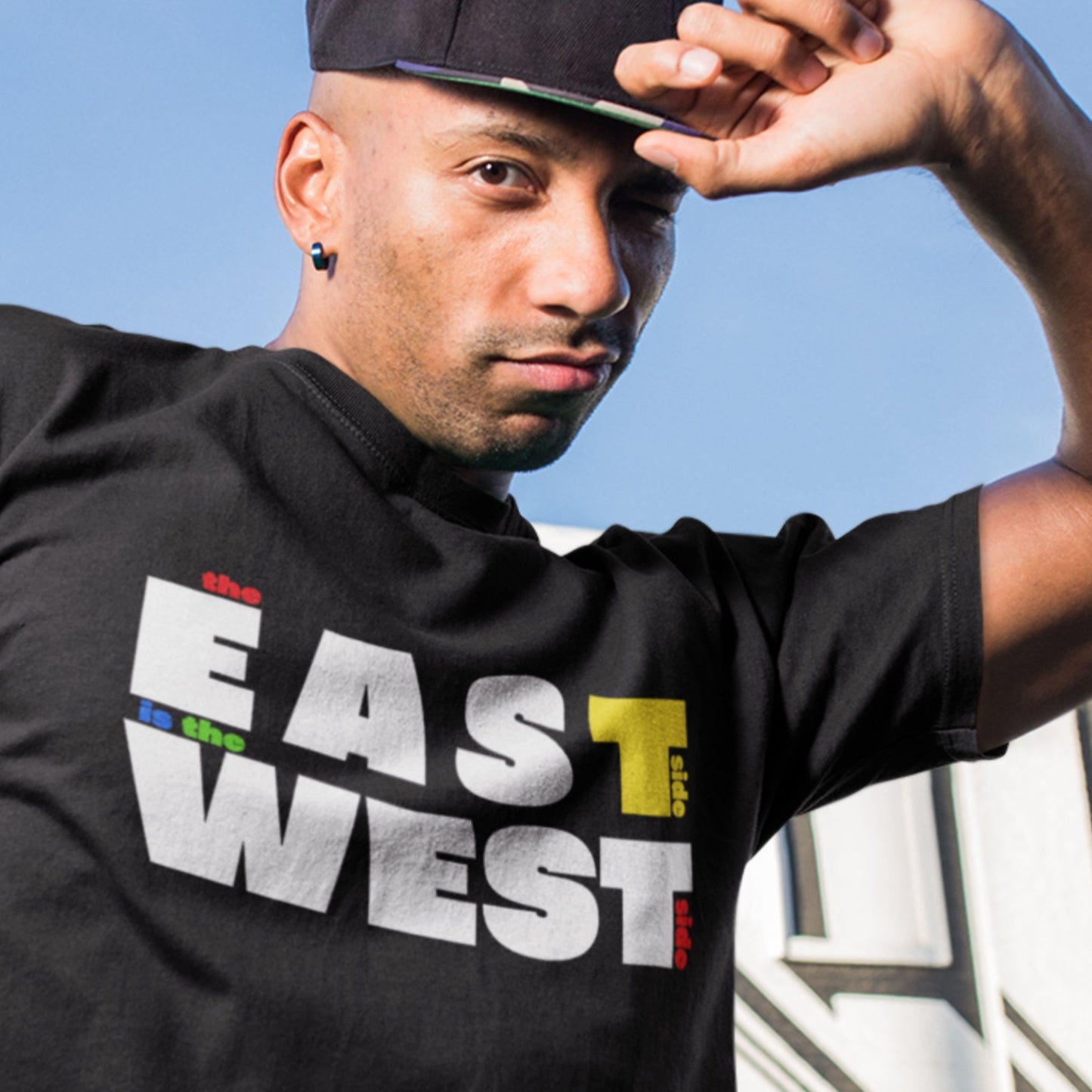 Cool guy wearing a black t-shirt with multicolored 'the east side is the west side' text outside.