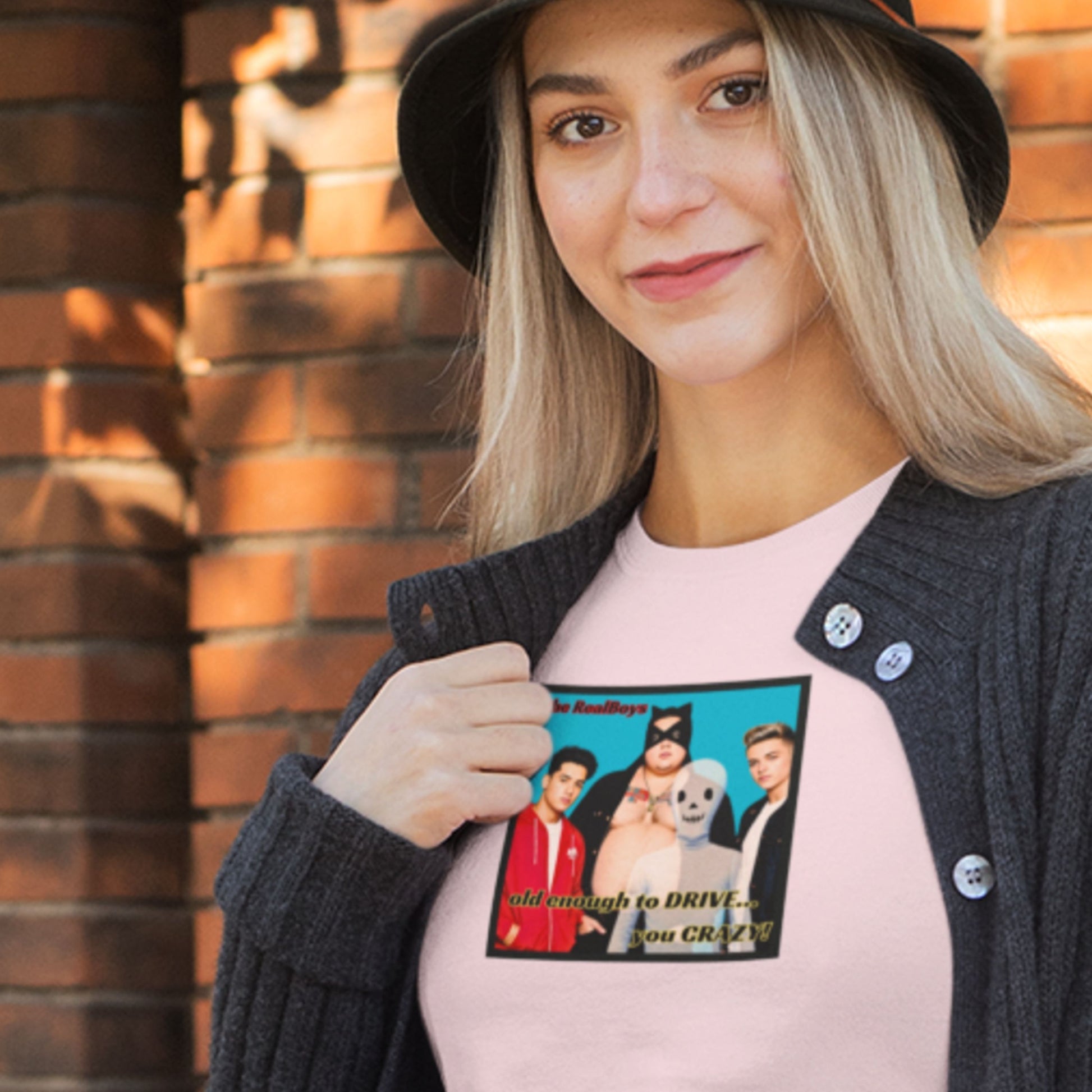 Hat woman wearing a pink t-shirt featuring a graphic of the debut album from the RealBoys in front of a brick wall.