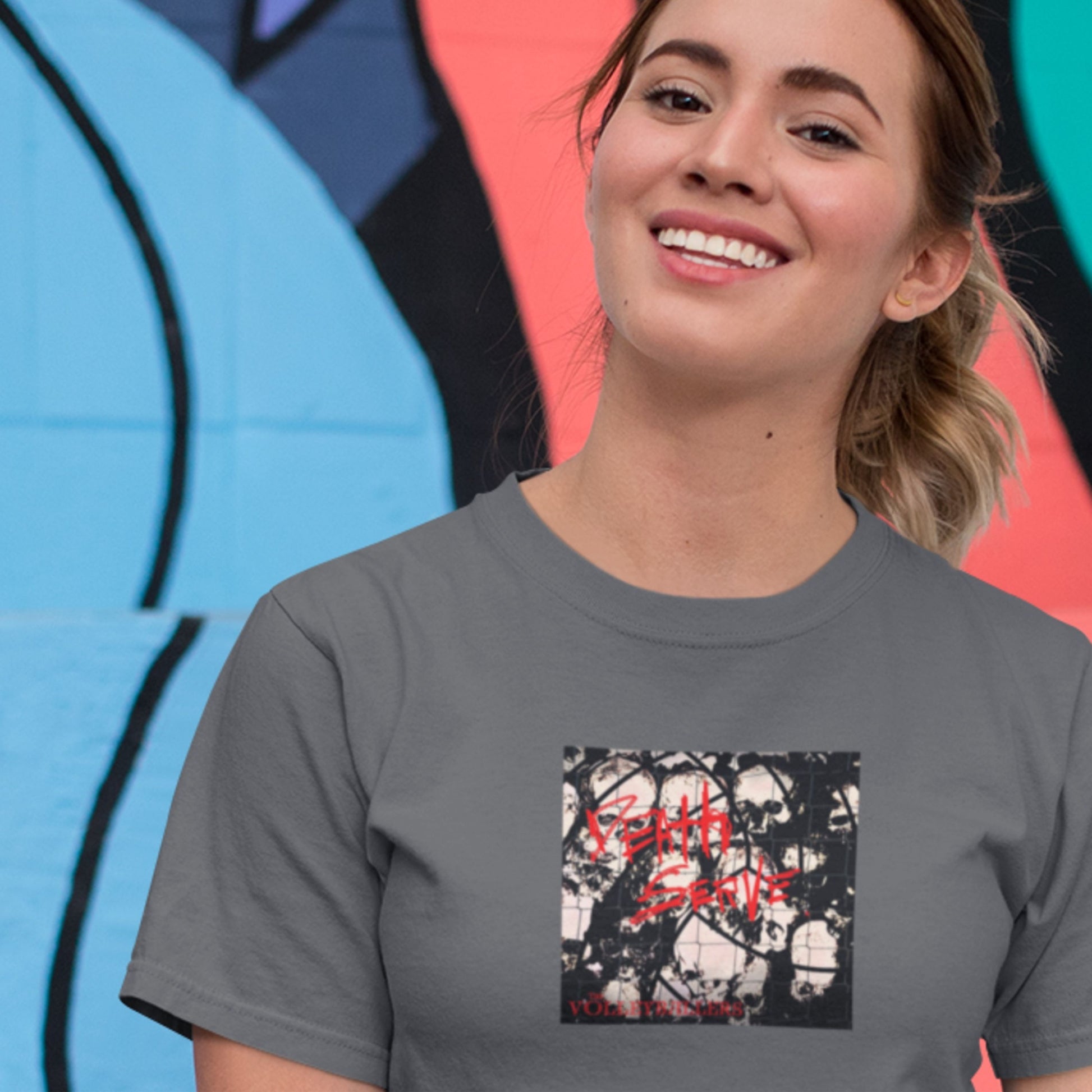 Woman wearing a t-shirt with an image of the Volleyballers album Death Serve in front of a colorful mural