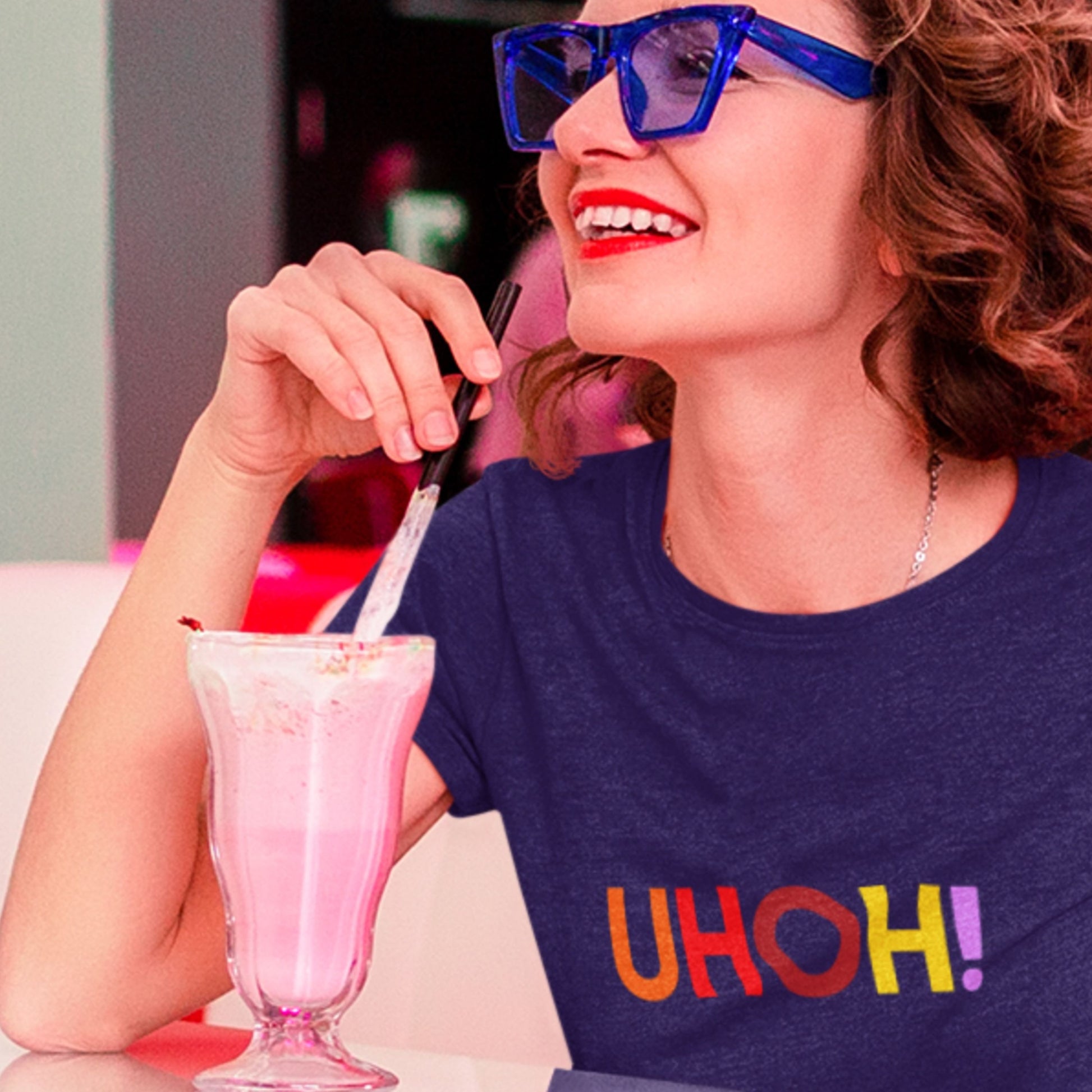 Woman wearing sunglasses and a 'UHOH!' shirt, enjoying a pink milkshake.