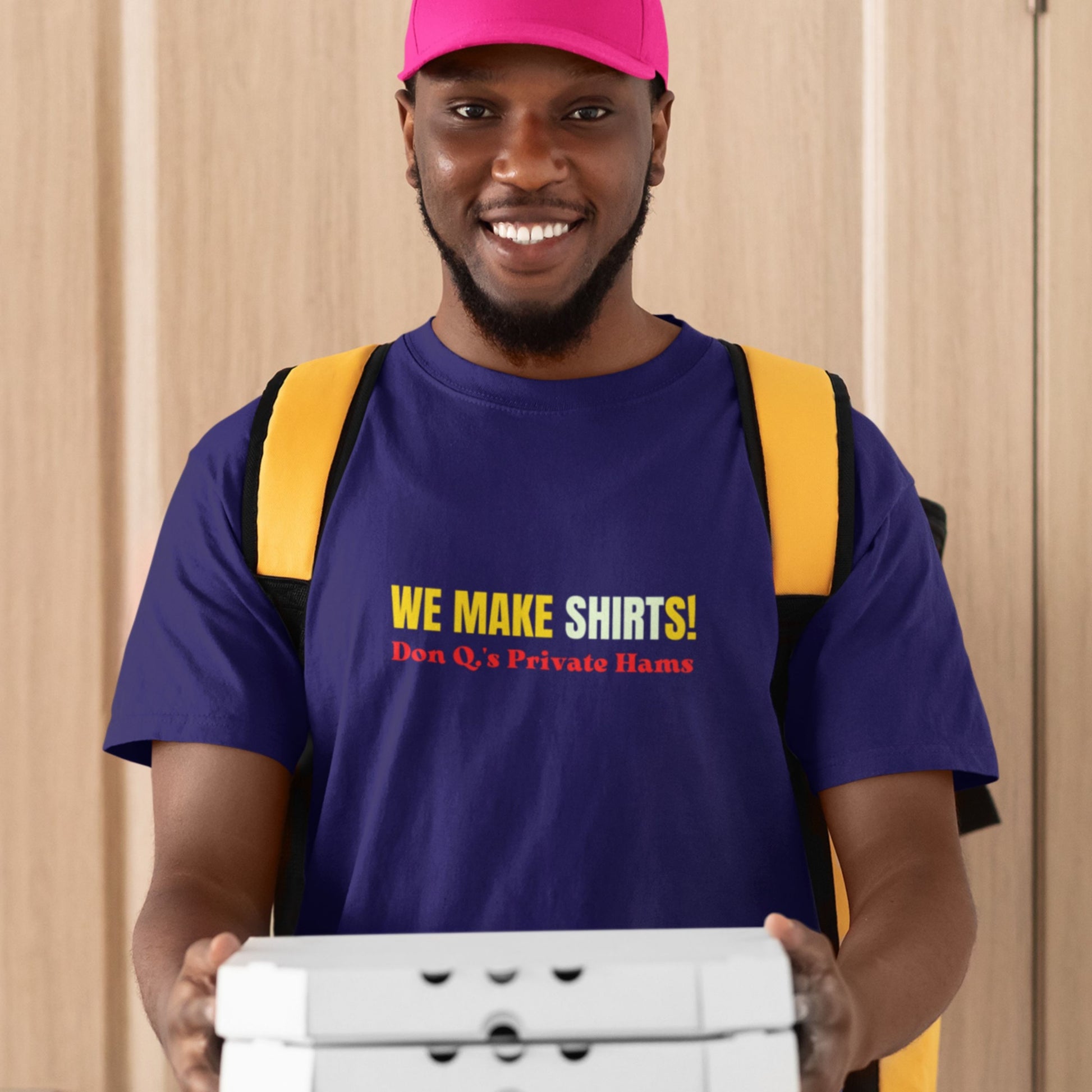 Man wearing a purple t-shirt with 'We Make Shirts!' text, holding a box against a beige curtain background.
