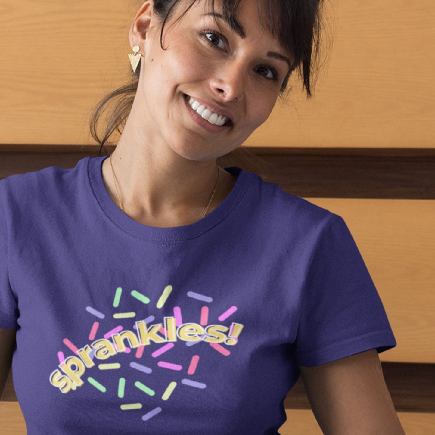 Woman wearing a purple t-shirt with 'sprankles!' design against a wooden background.