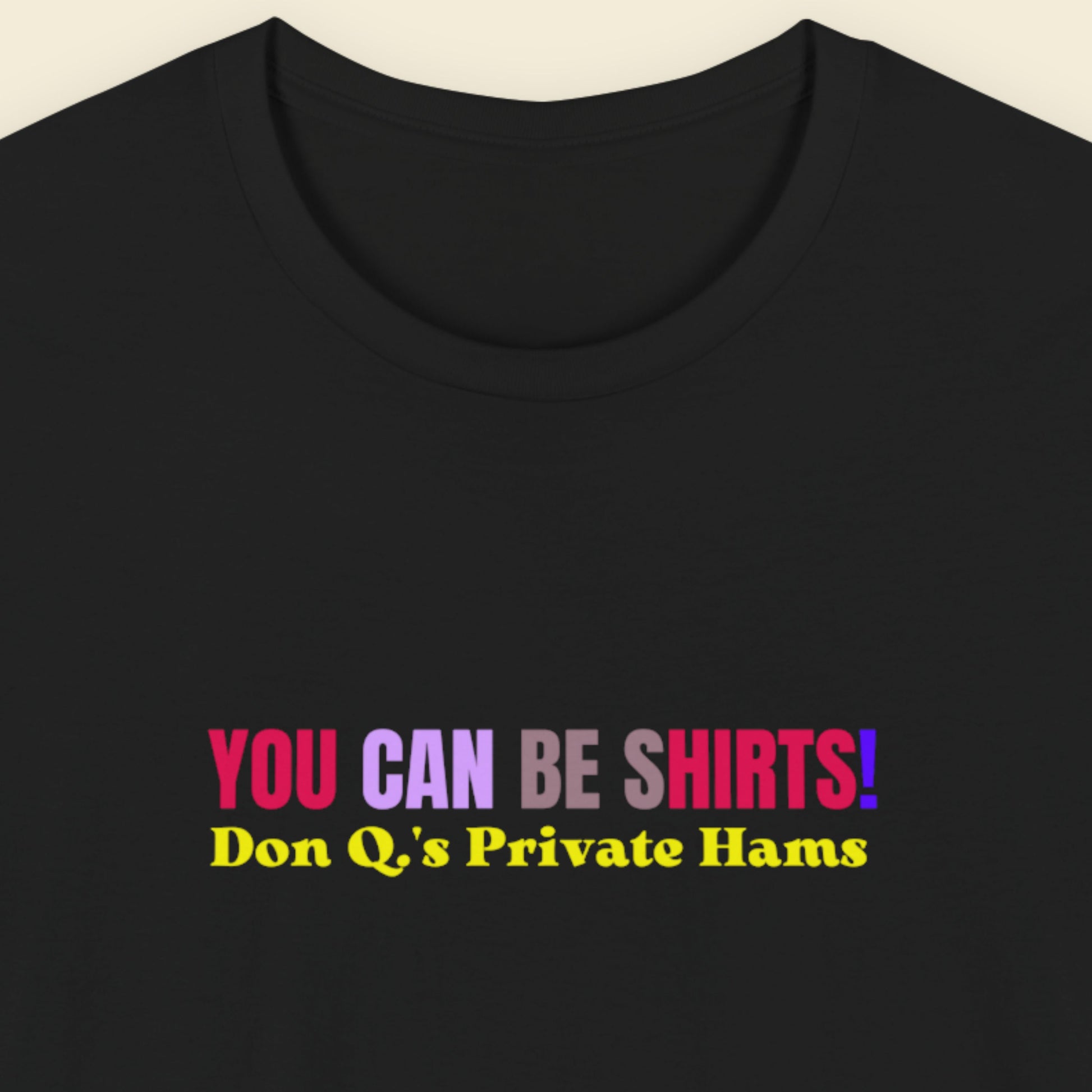 Black t-shirt with multicolored 'you can be shirts!' text on a beige background.