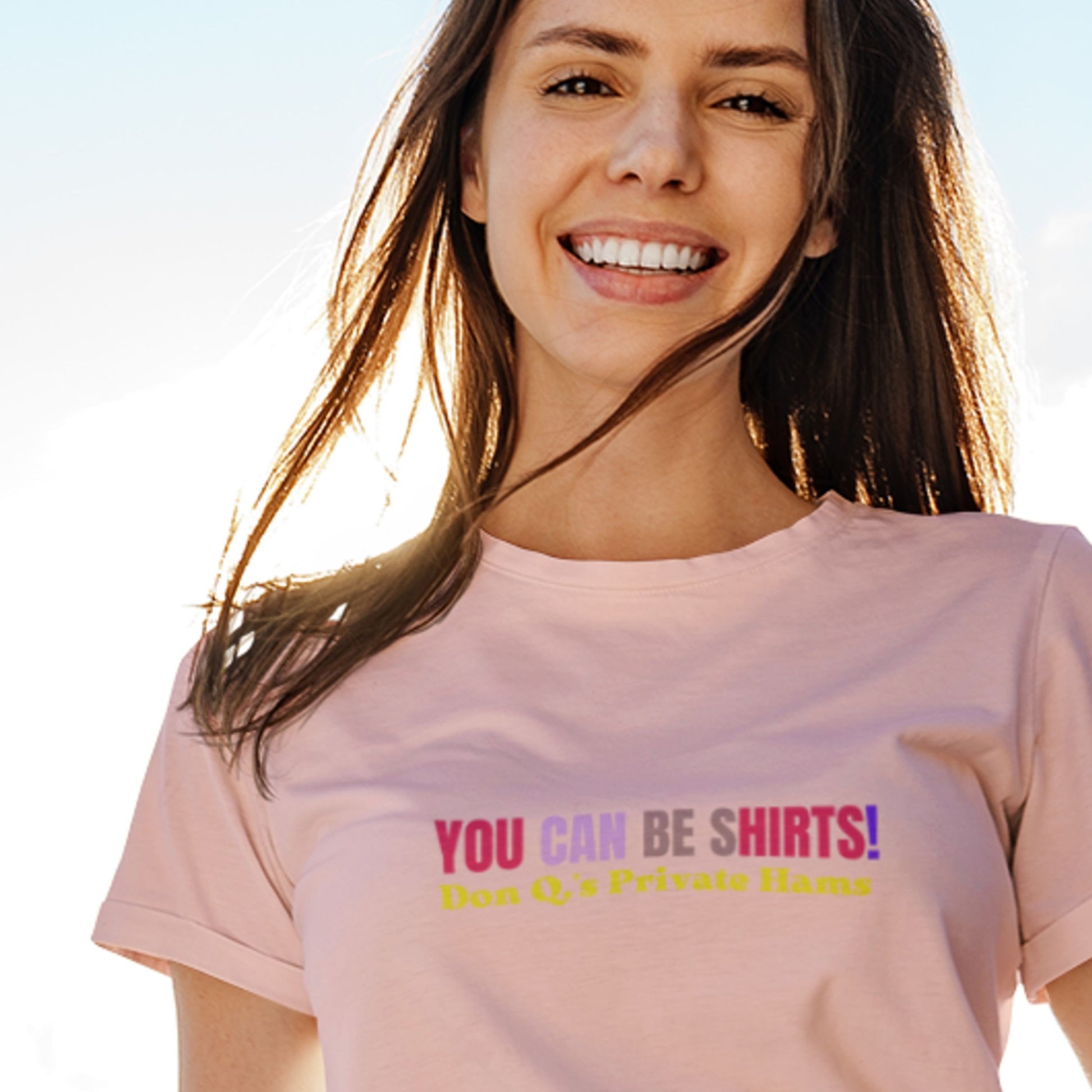 Smiling woman wearing a pink t-shirt with multicolored 'you can be shirts!' text against a clear sky.