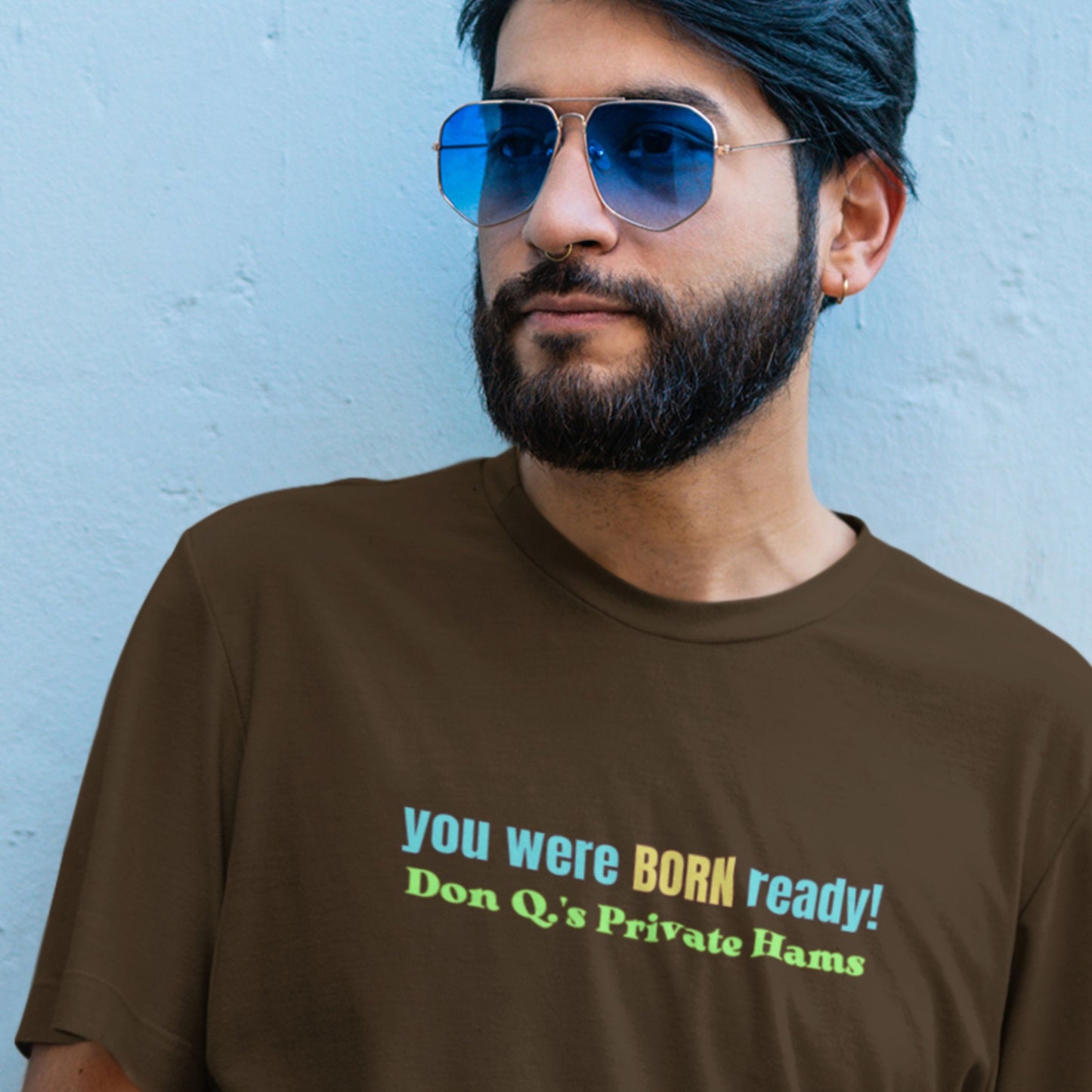 Man wearing sunglasses and a brown t-shirt with humorous text against a light blue wall.